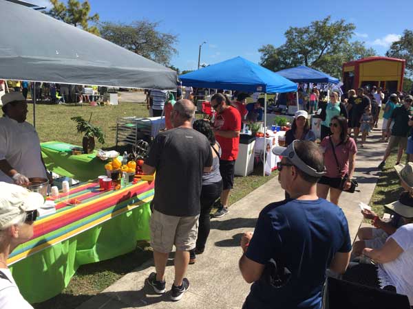 Crowd of people gathered around outdoor food stalls with colorful tablecloths and canopy tents on a sunny day.